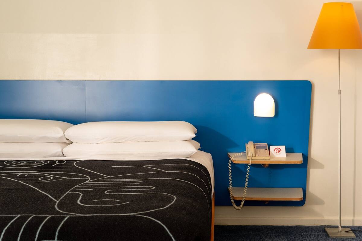 A hotel room with a blue headboard, a bedside table with a phone and a card, and a lamp.
