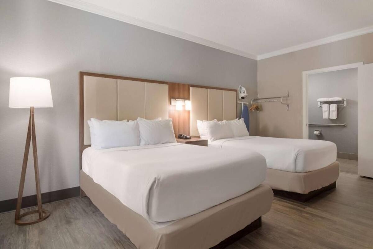 Ein Hotelzimmer mit zwei Betten, einem Nachttisch mit Lampe, einem Spiegel und einem Badezimmer mit Handtuchhalter.