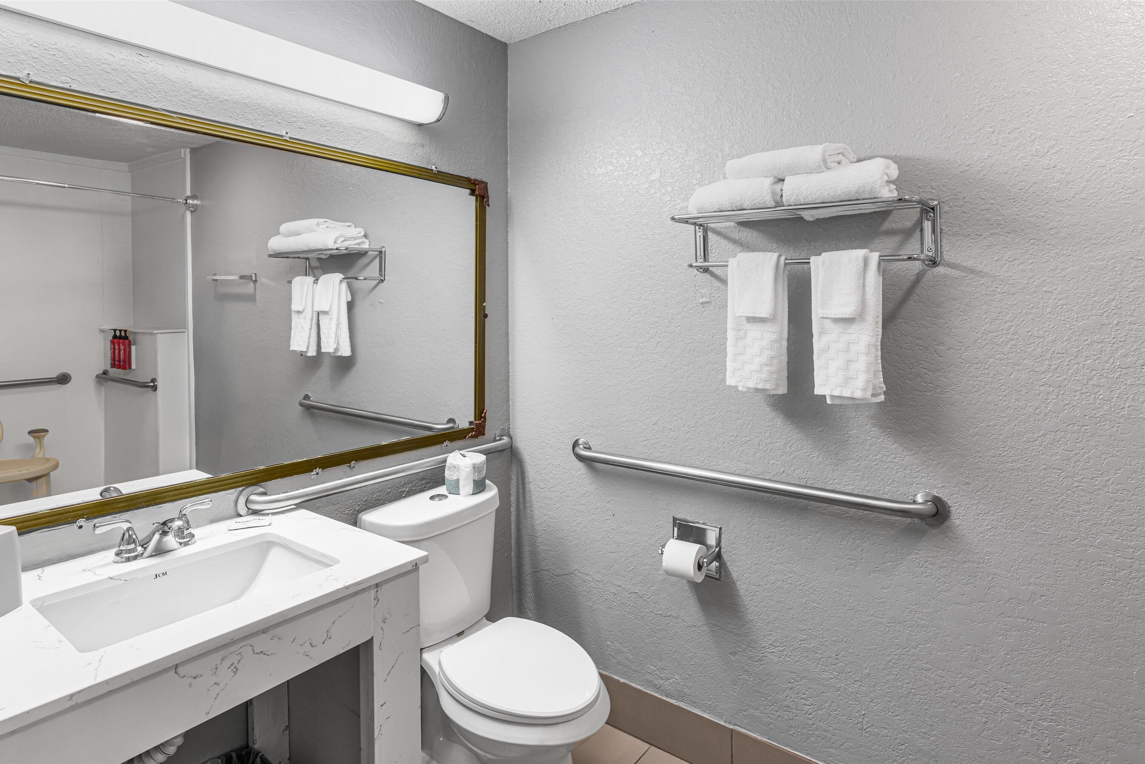 Une salle de bain moderne avec un lavabo, des toilettes et un miroir, tous de couleur blanche. On y trouve des porte-serviettes au mur et une barre d’appui près des toilettes.