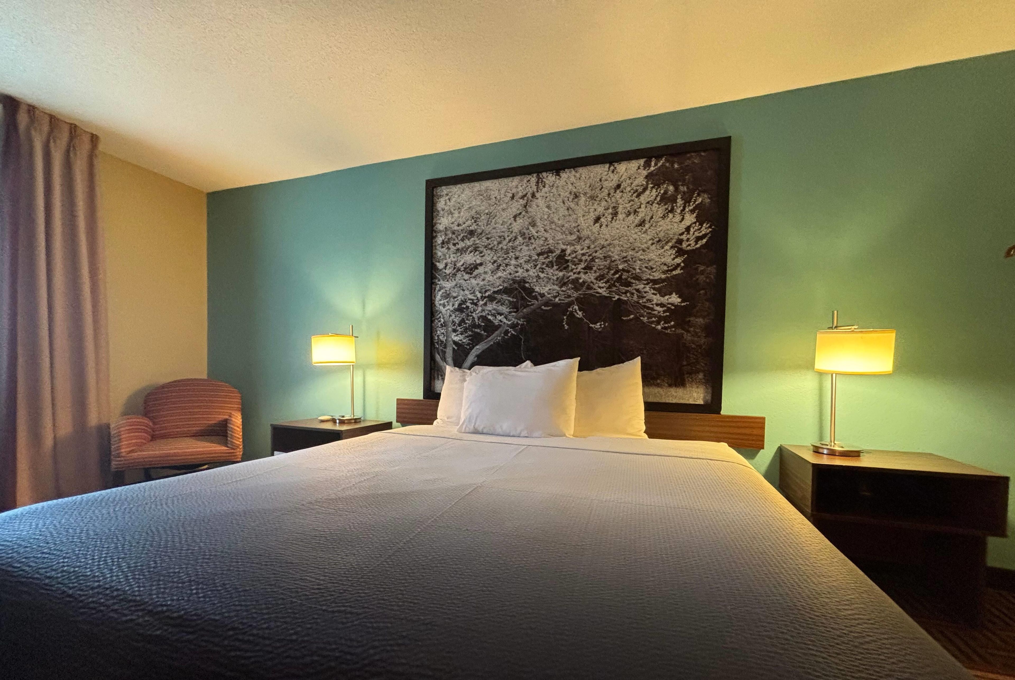 A hotel room with a large bed, two bedside tables with lamps, a chair, and a large framed picture on the wall.