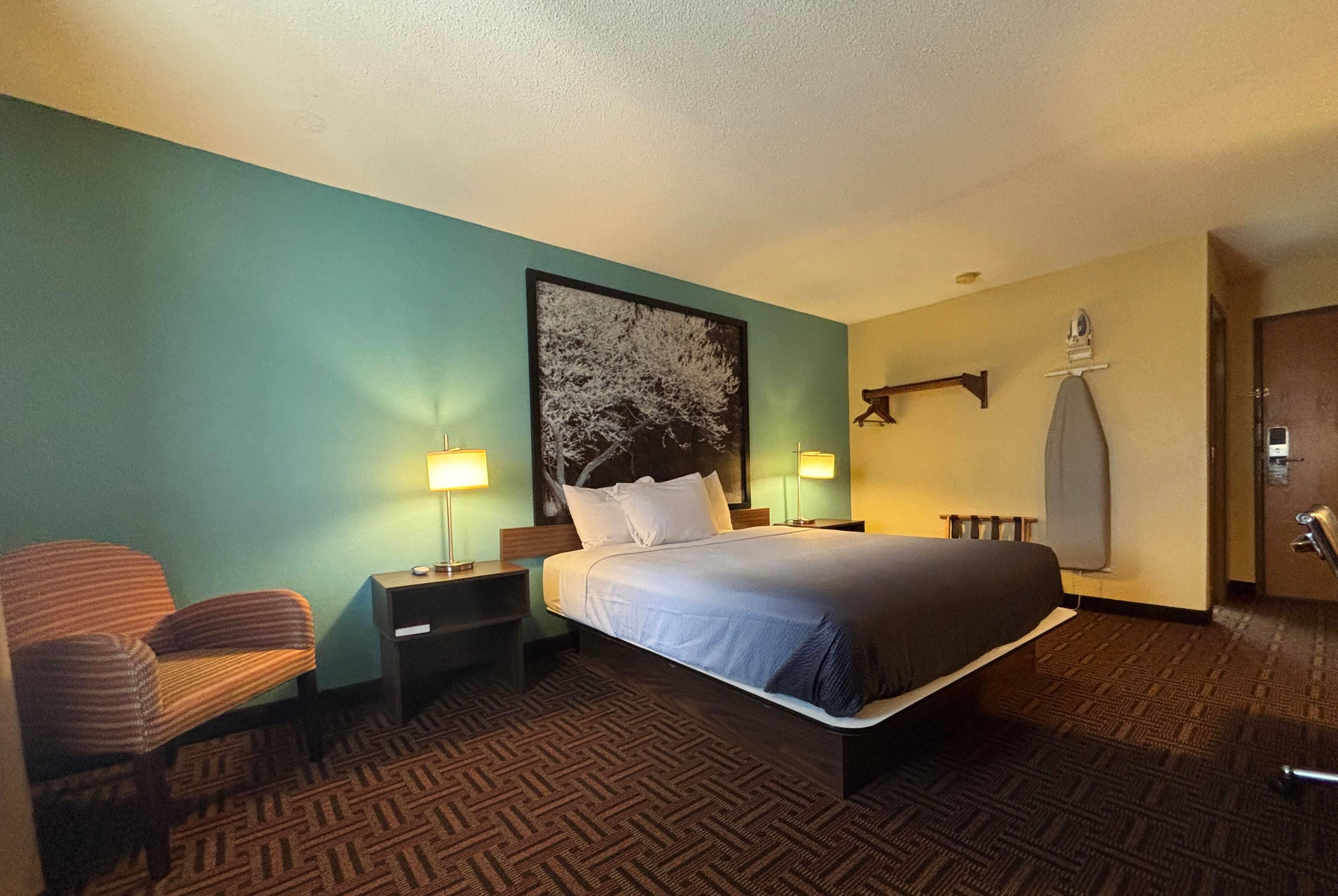 A hotel room with a bed, two bedside tables with lamps, a chair, a wall-mounted towel rack, and a door.