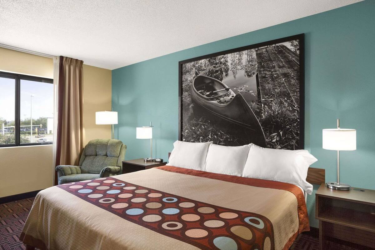 A hotel room with a bed, two bedside lamps, a chair, a large window, and a wall with a black and white photograph.