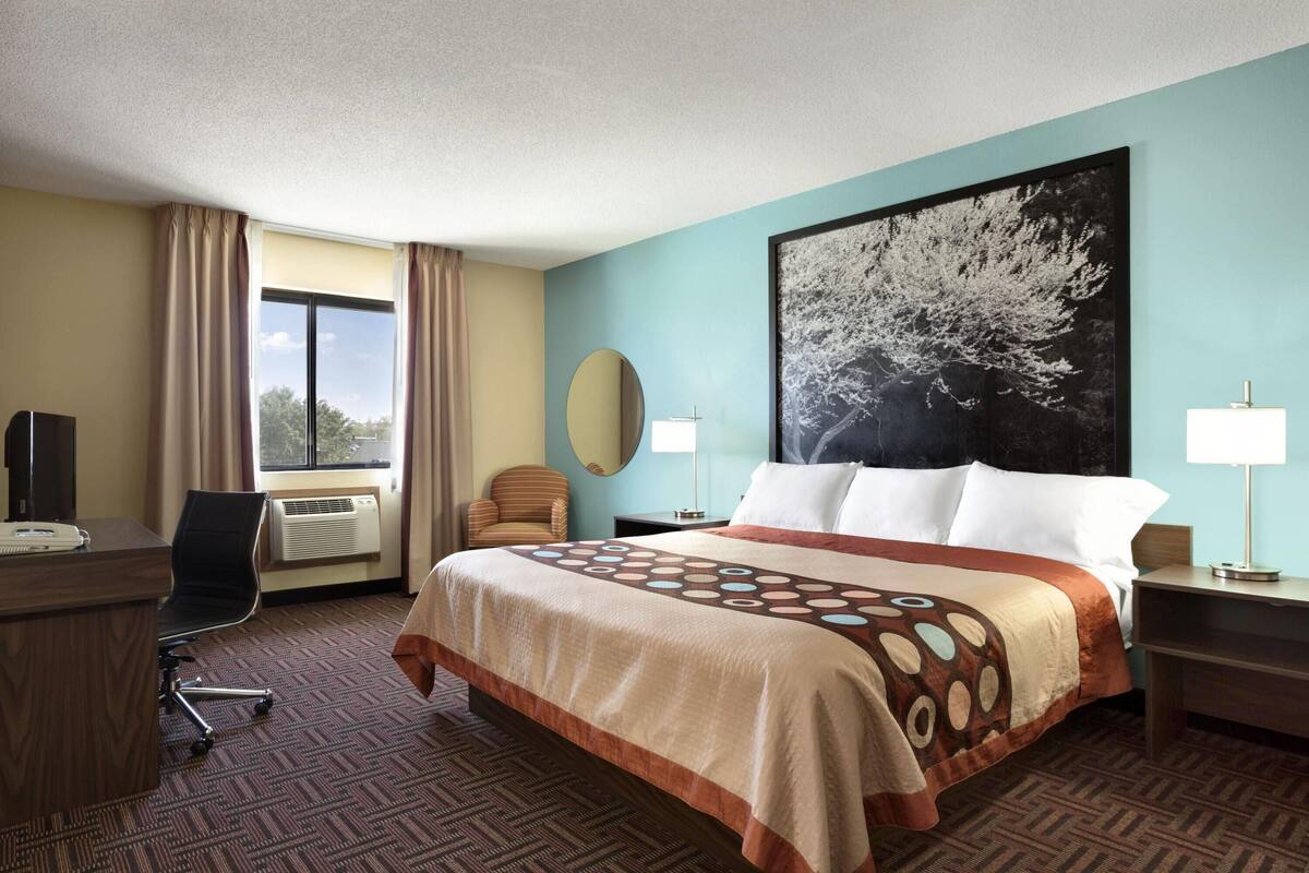A hotel room with a large bed, a desk, a chair, a window with curtains, a wall-mounted TV, and a large painting.