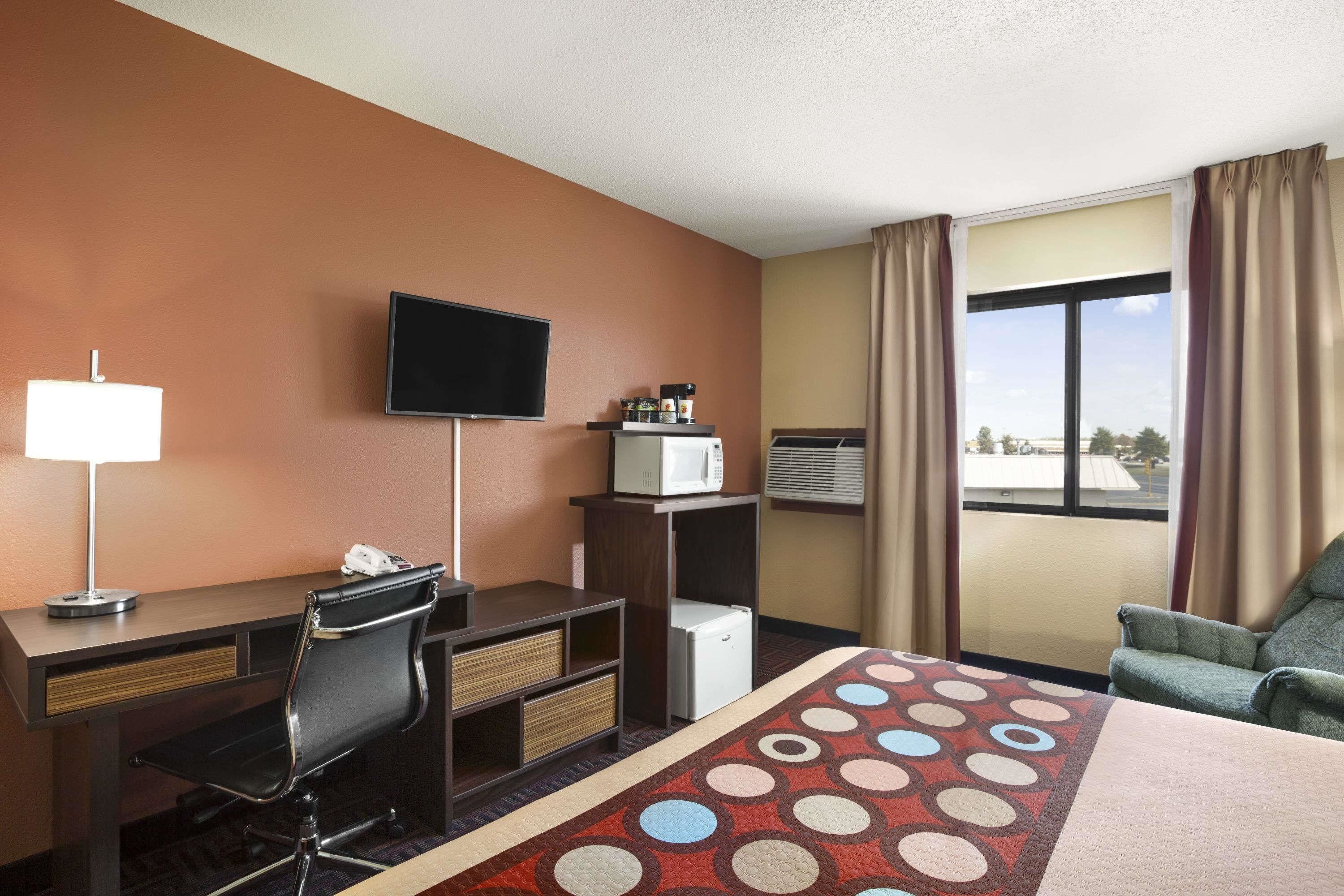 A hotel room with a desk, chair, TV, microwave, and a view of the outdoors.