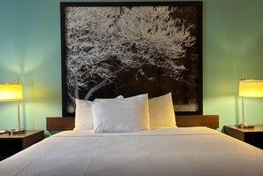 Een hotelkamer met een groot schilderij van een boom aan de muur, een opgemaakt bed met witte lakens en twee bedlampjes.
