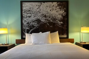 Habitación de hotel con un cuadro grande de un árbol en la pared, una cama bien hecha con ropa blanca y dos lámparas de noche.