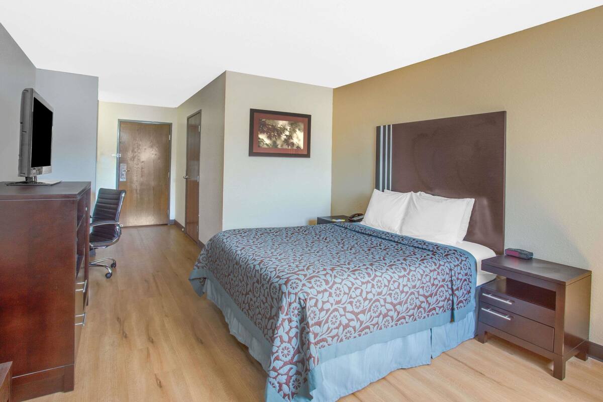 A hotel room with a bed, a desk with a computer, a chair, a nightstand, and a framed picture on the wall.