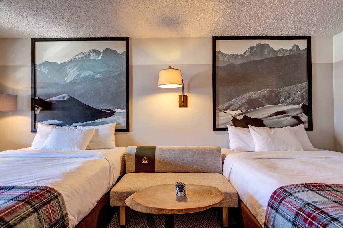 Une chambre d’hôtel avec deux lits, une petite table ronde, une lampe et des tableaux représentant des paysages de montagne accrochés aux murs.