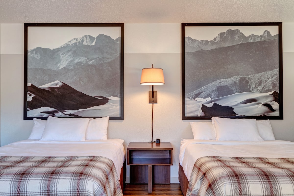 Une chambre d’hôtel avec deux lits, une table de chevet avec une lampe et deux tableaux encadrés représentant des paysages de montagne accrochés au mur.