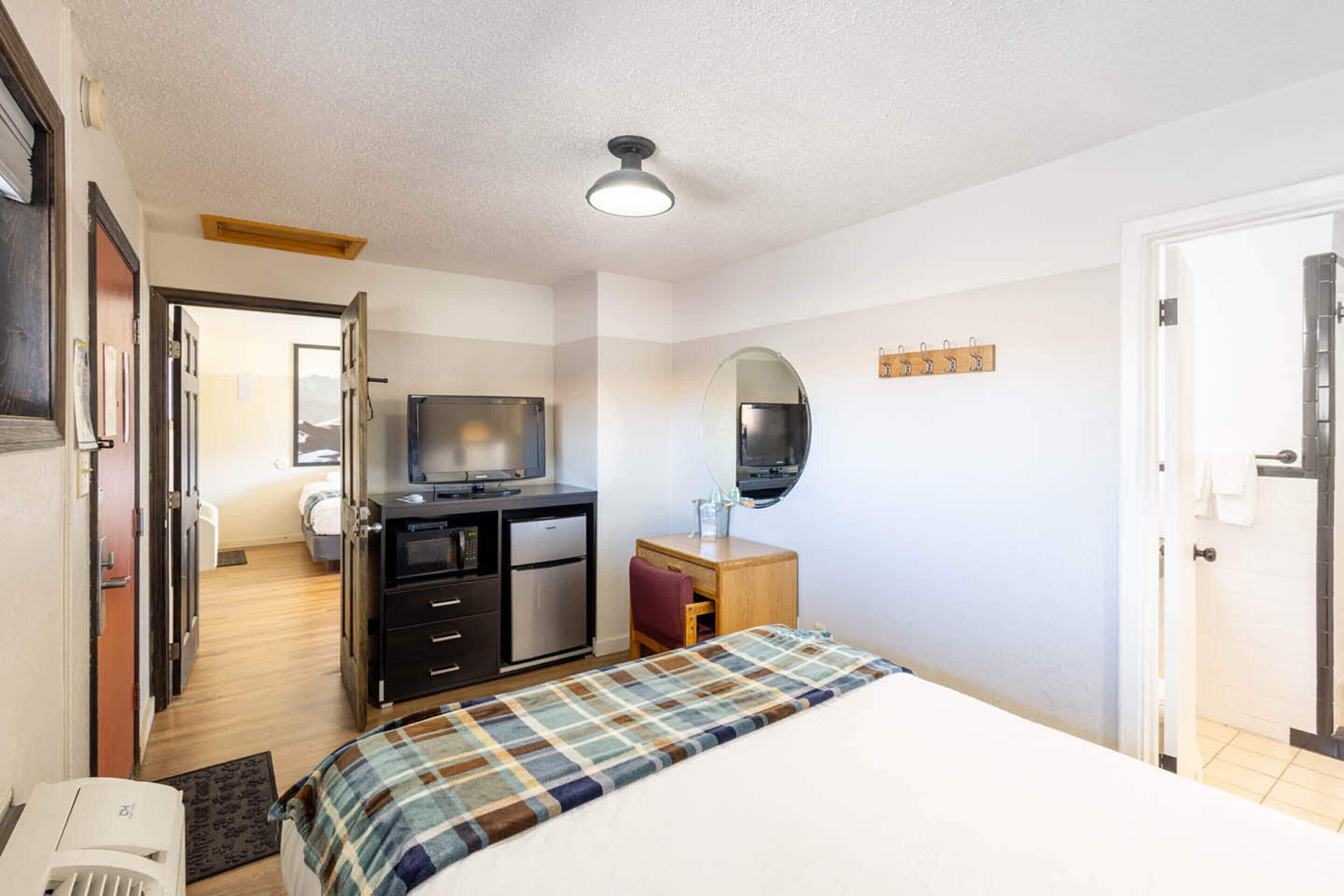 Una habitación de hotel con cama, escritorio, espejo, televisor y kitchenette.