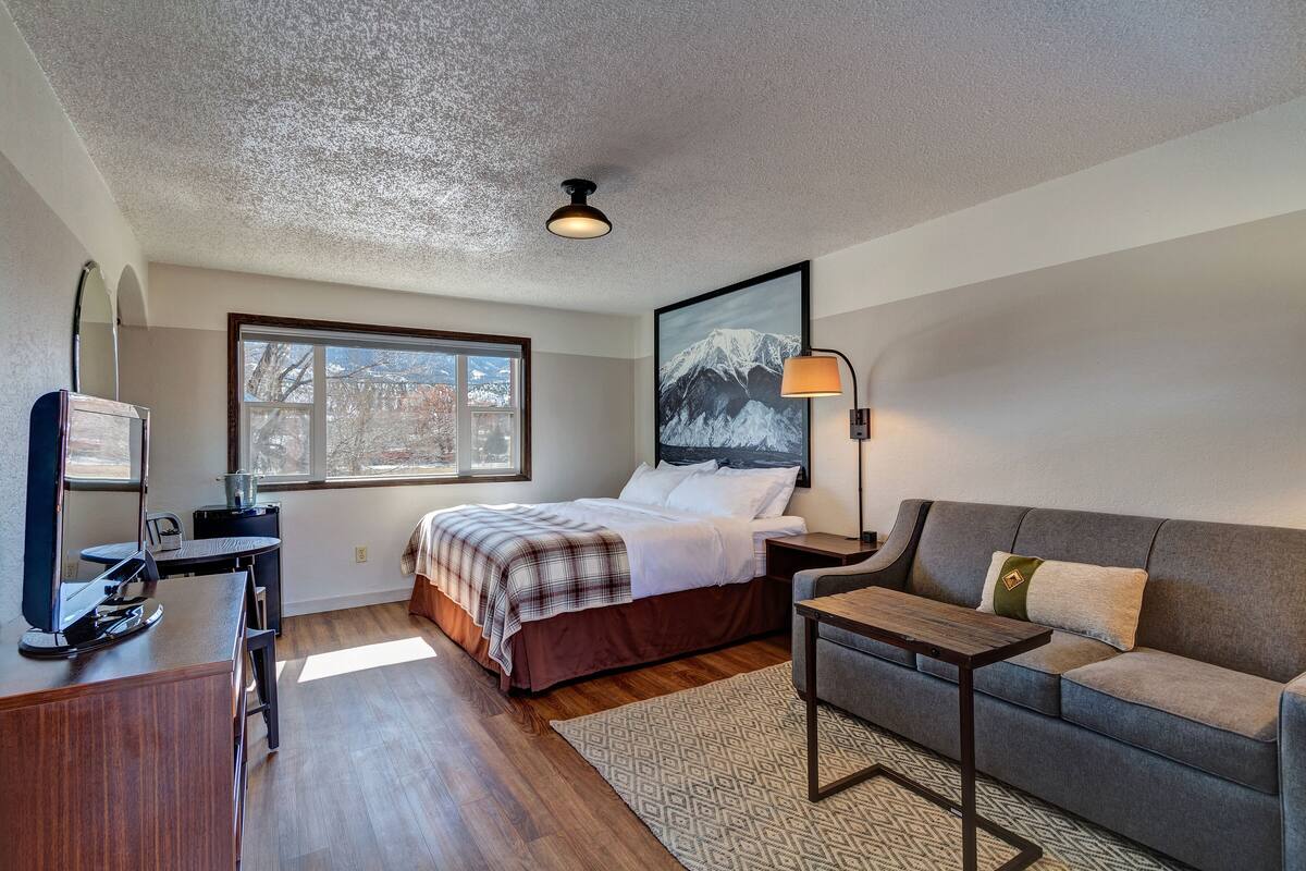 Une chambre à coucher avec un lit, un bureau avec un ordinateur, un canapé, une lampe et une fenêtre donnant sur les montagnes.