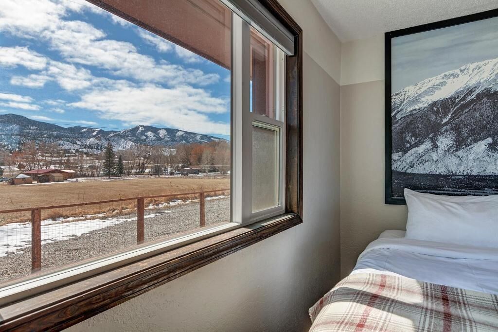 A bedroom with a large window, a bed with white bedding, and a framed picture of a snowy mountain landscape.