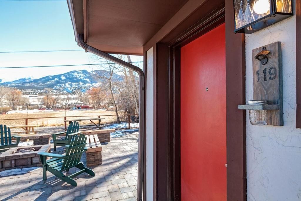 Una puerta roja con placa de número de casa, mobiliario exterior y vistas a un paisaje nevado.
