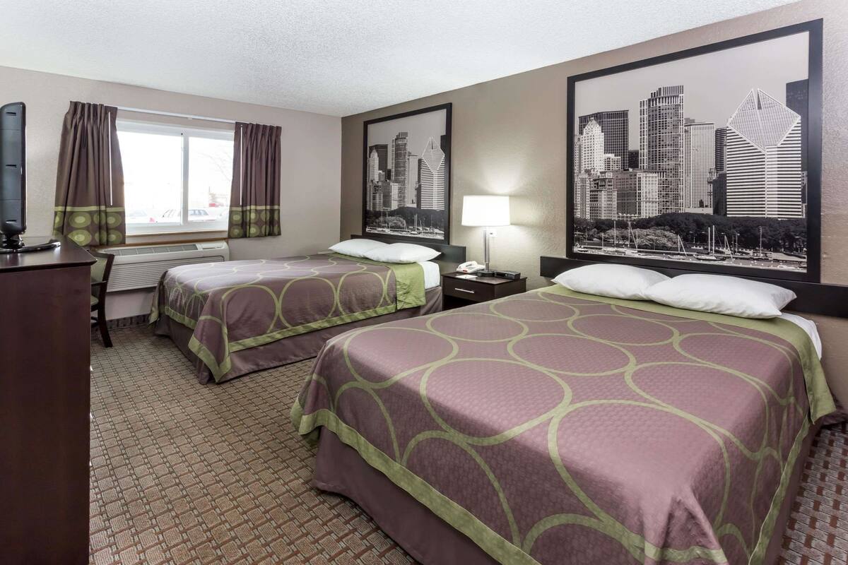 A hotel room with two beds, a desk, a chair, a TV, a window with curtains, and a cityscape painting on the wall.