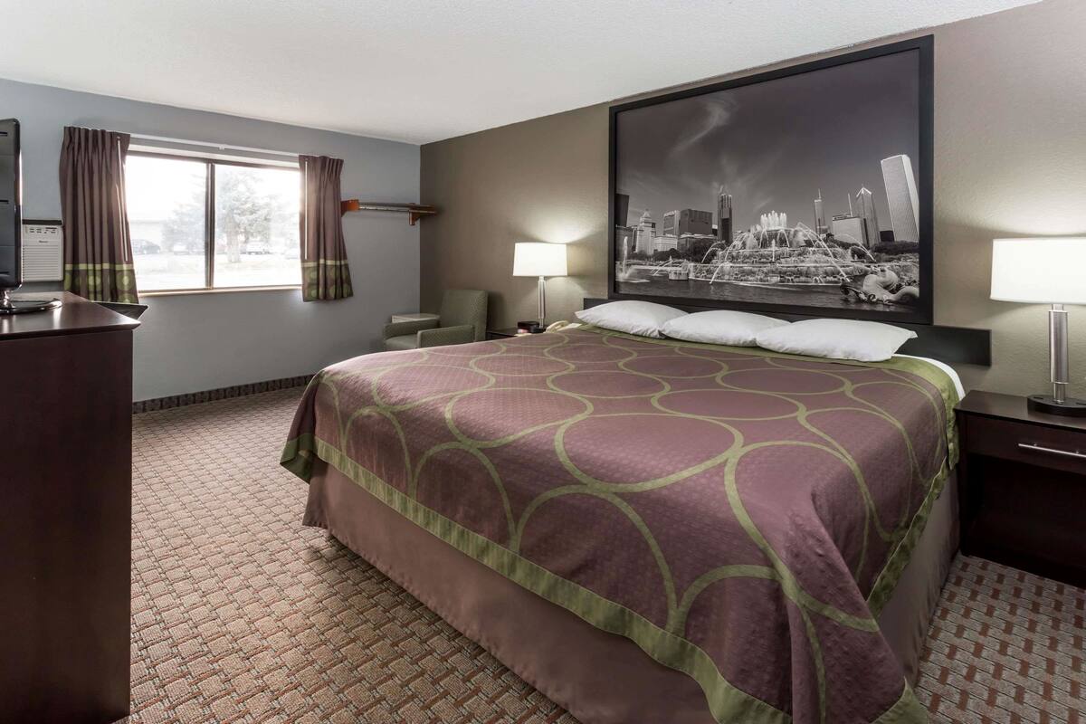 A hotel room with a large bed, a desk, a chair, a window with curtains, a cityscape painting, and a lamp.