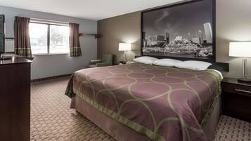 A hotel room with a large bed, a desk, a chair, a window with curtains, a cityscape painting, and a lamp.