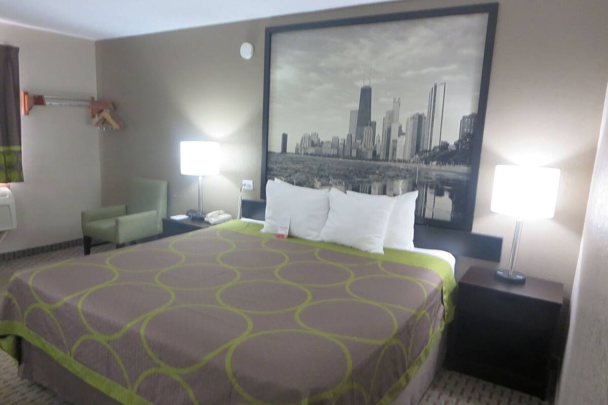 A hotel room with a large bed, two bedside tables, a chair, a desk, a lamp, and a cityscape painting on the wall.