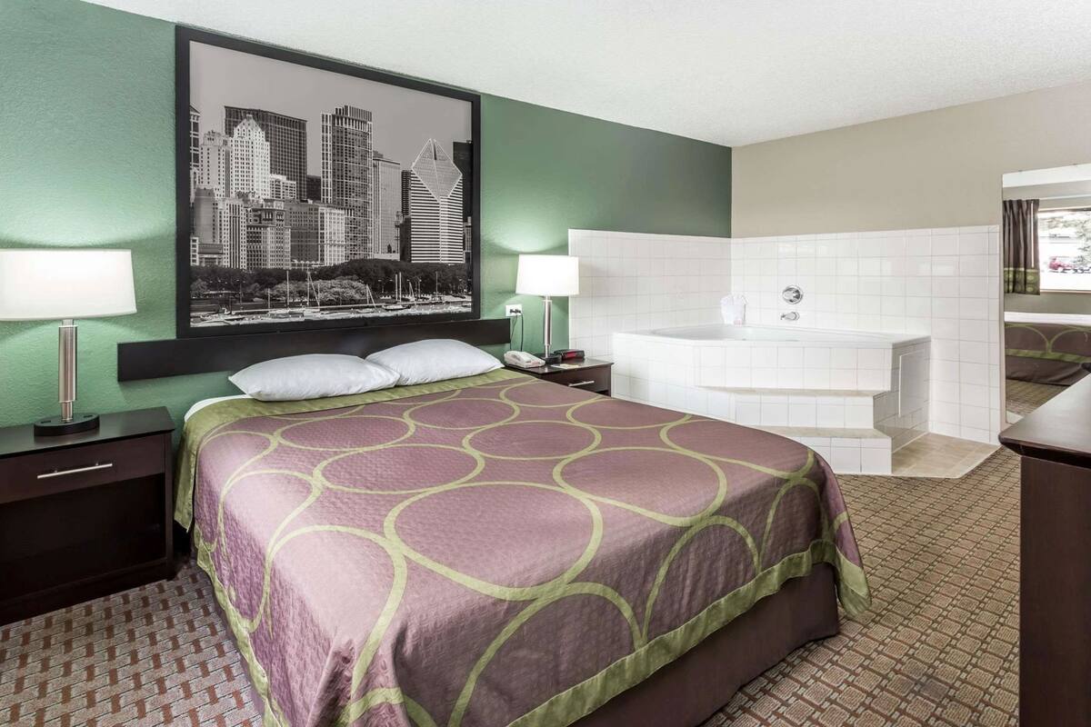 A hotel room with a bed, two bedside lamps, a cityscape artwork, a large bathtub, and a view of the outside.