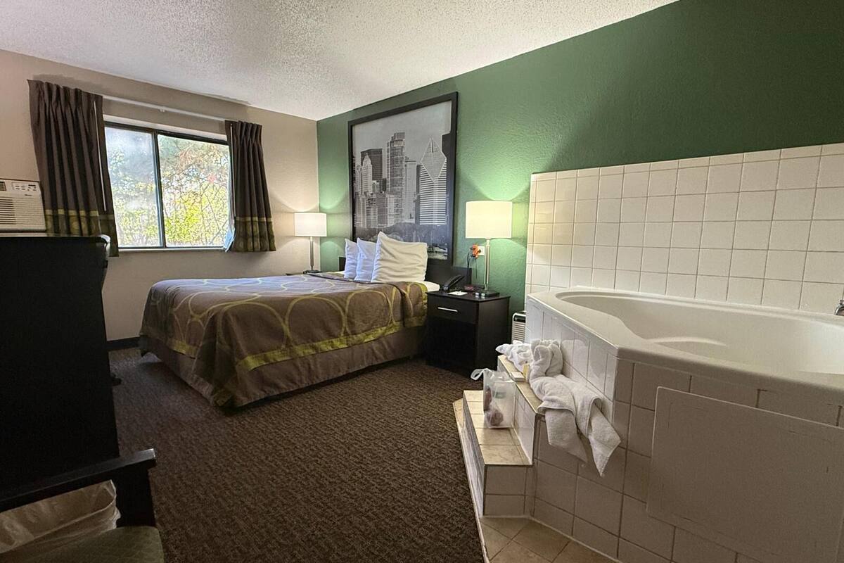 A hotel room with a bed, a desk, a chair, a window with curtains, a painting, a lamp, and a bathtub with towels.