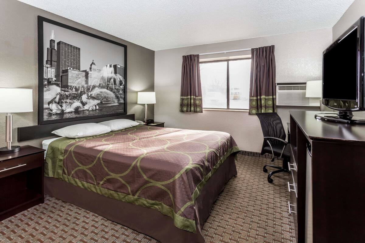 A hotel room with a large bed, a desk with a computer, a chair, a TV, a window with curtains, and a wall-mounted air conditioner.