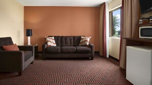 A hotel room with a dark brown sofa, a smaller armchair, a flat-screen TV, a mini-fridge, and a large window with curtains.