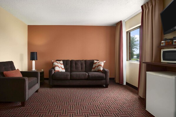 A hotel room with a dark brown sofa, a smaller armchair, a flat-screen TV, a mini-fridge, and a large window with curtains.
