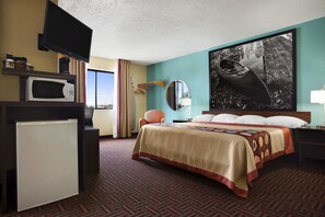 A hotel room with a bed, a TV, a microwave, a refrigerator, a large window, and a painting on the wall.