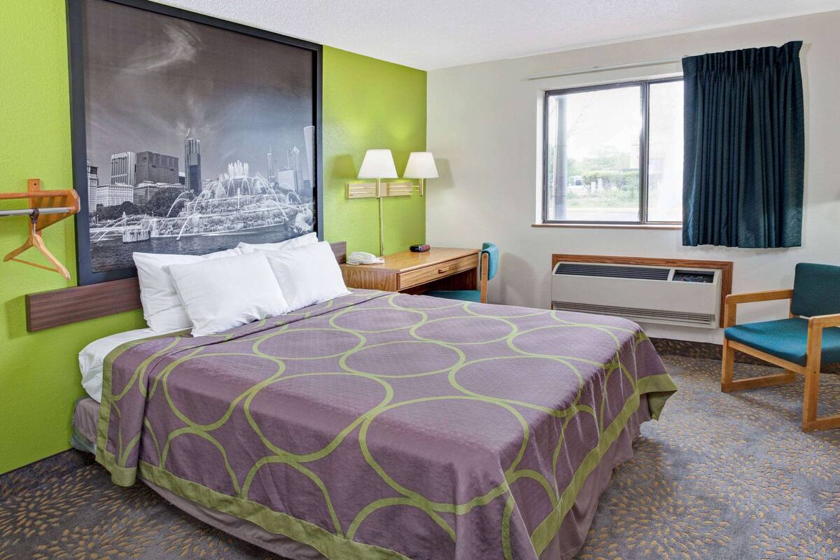 A hotel room with a bed, a desk, a chair, a window with curtains, and a wall-mounted air conditioner.