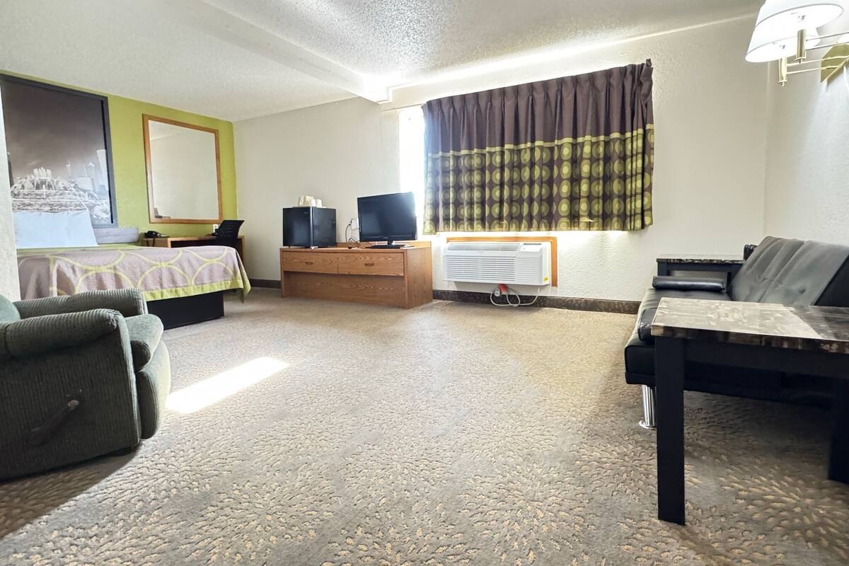 A hotel room with a bed, a desk with two monitors, a chair, a sofa, a lamp, and an air conditioning unit.