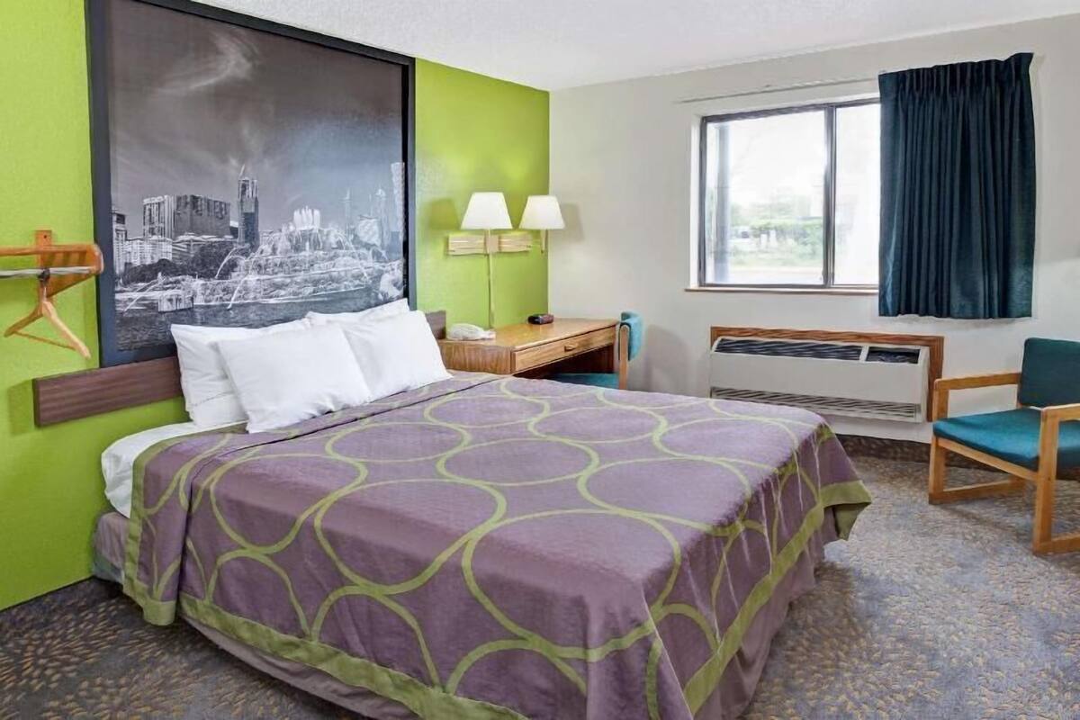 A hotel room with a bed, a desk, a chair, a window with curtains, a wall-mounted air conditioner, and a cityscape painting.