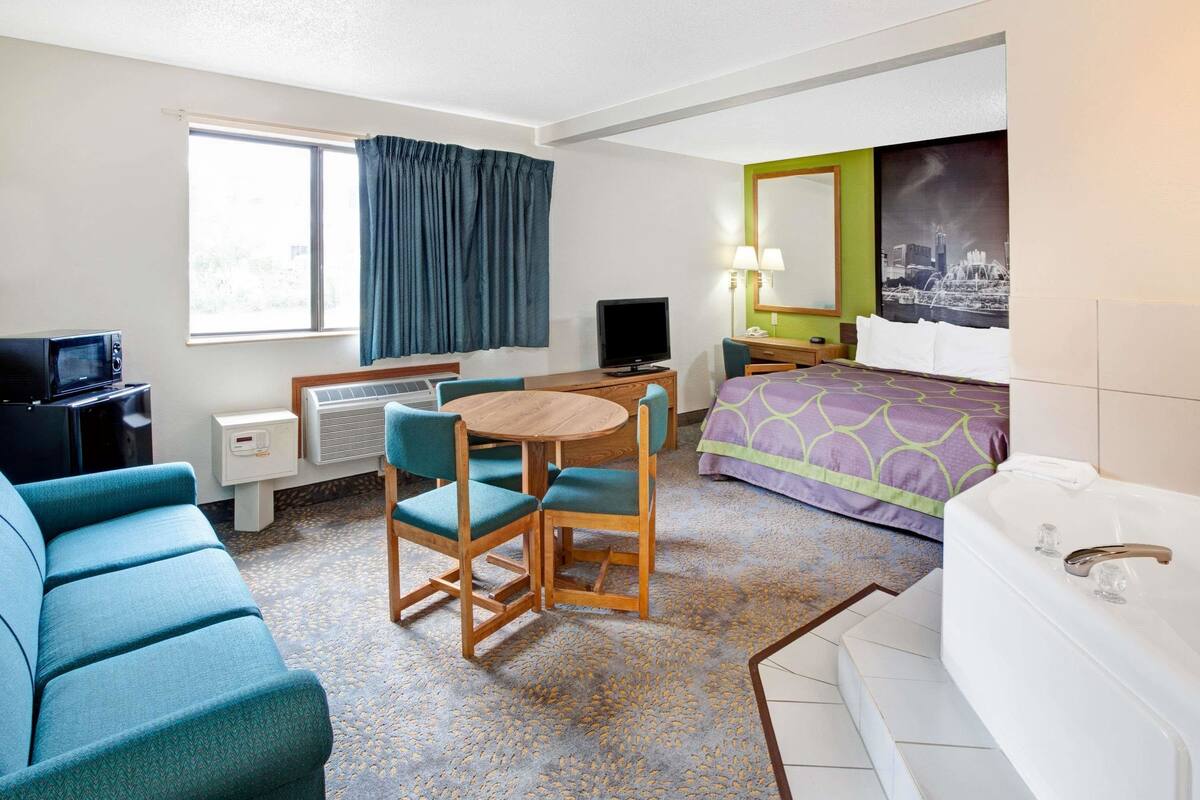 A hotel room with a bed, a desk with a computer, a sofa, a chair, a TV, a window with curtains, and a bathroom with a bathtub.