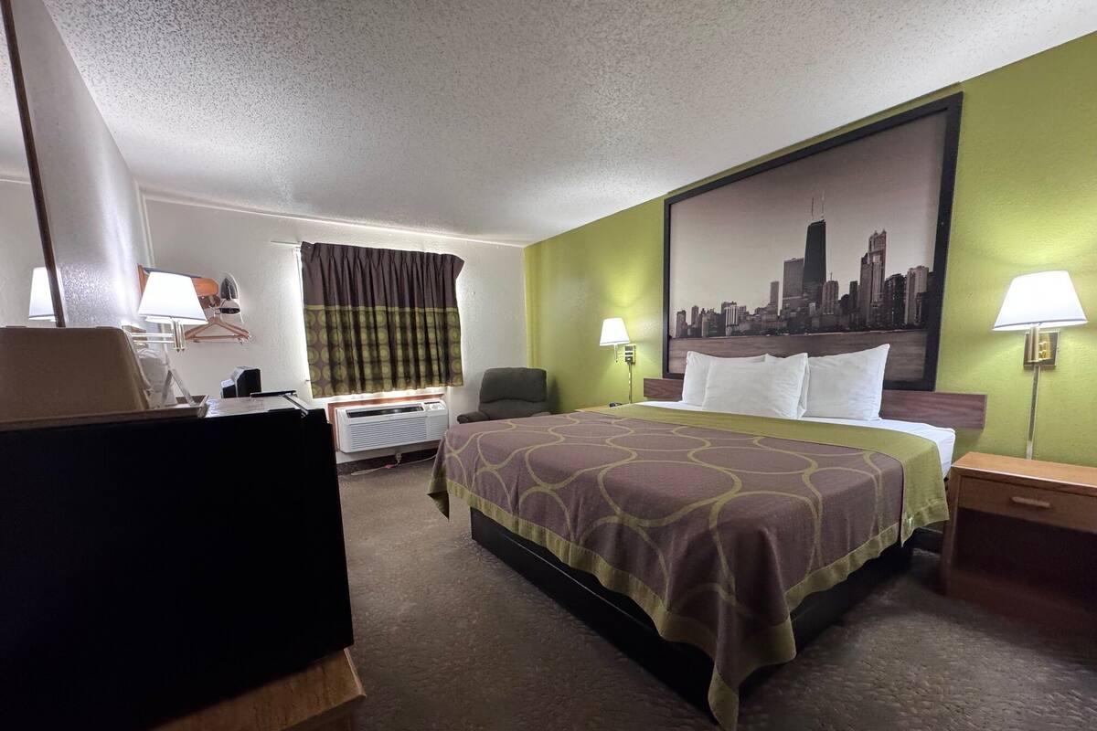 A hotel room with a bed, a desk, a chair, a window with curtains, a wall-mounted air conditioner, and a cityscape painting.