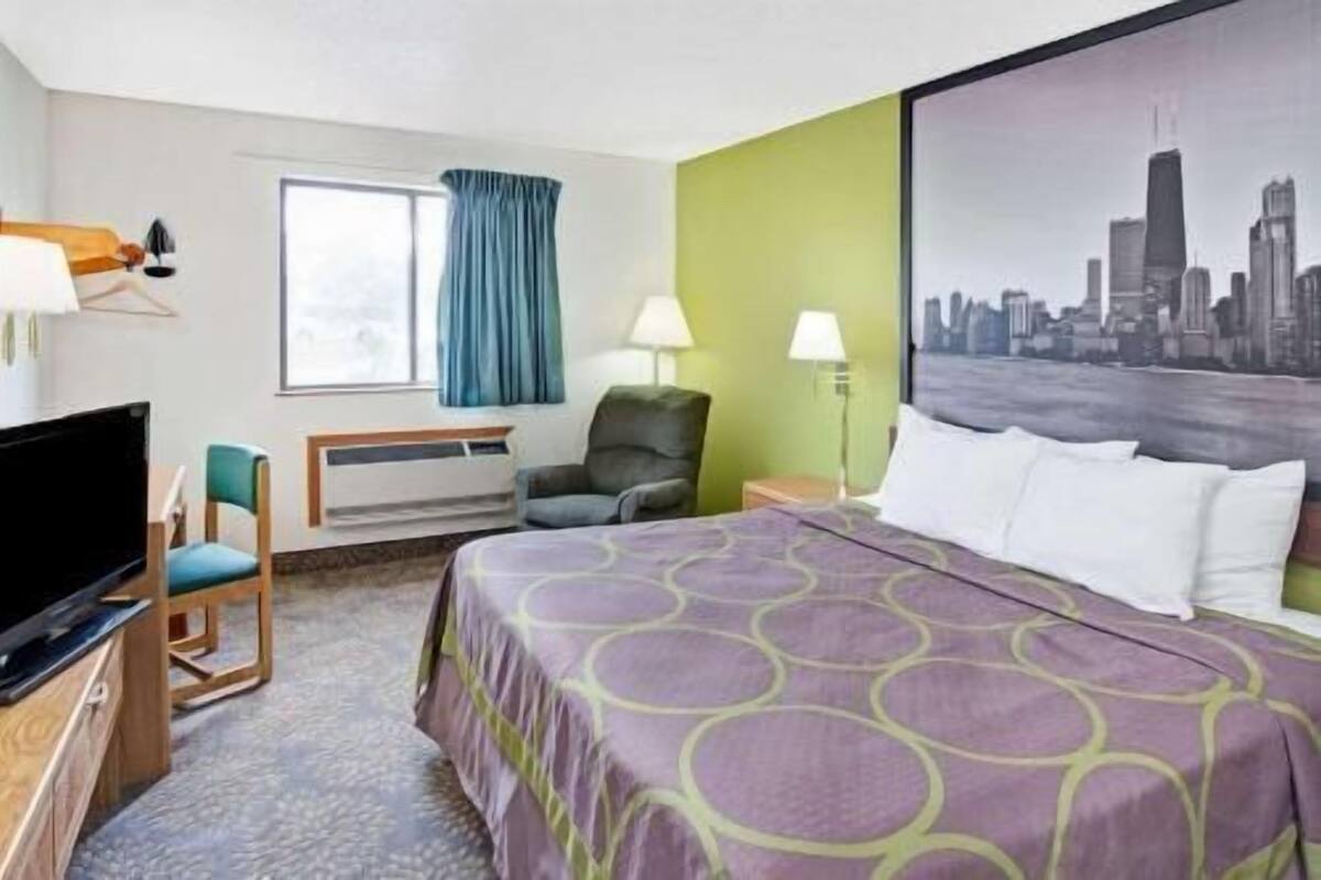 A hotel room with a bed, a chair, a desk with a TV, a window with curtains, and a cityscape view.