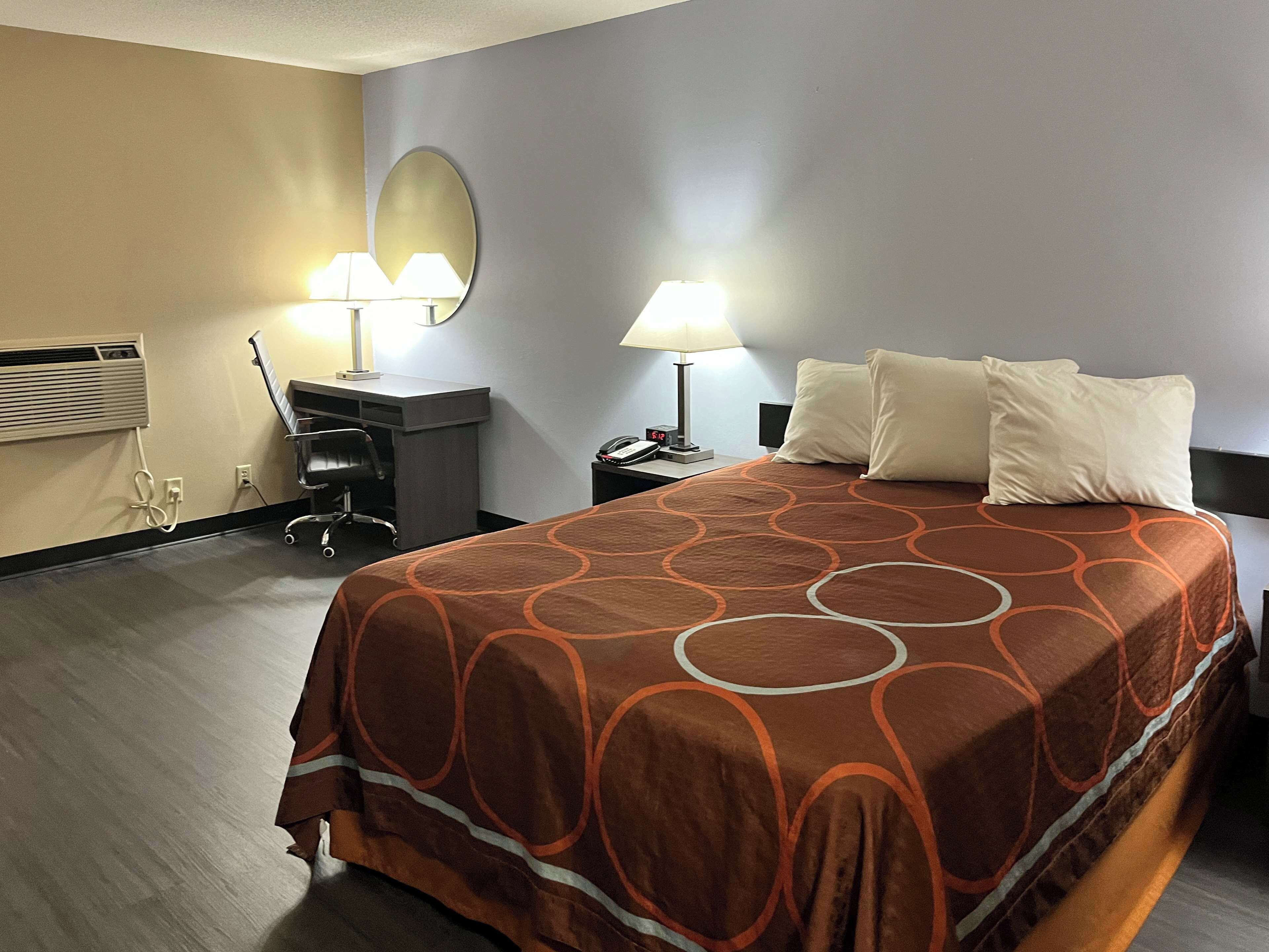 A hotel room with a bed, two bedside lamps, a desk with a chair, a mirror, and a wall-mounted heater.