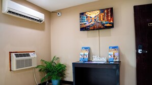 A reception desk with a 'Welcome' sign, a mounted air conditioner, a potted plant, and a TV displaying a bar scene. - Motel 6 Litchfield, IL (Litchfield)