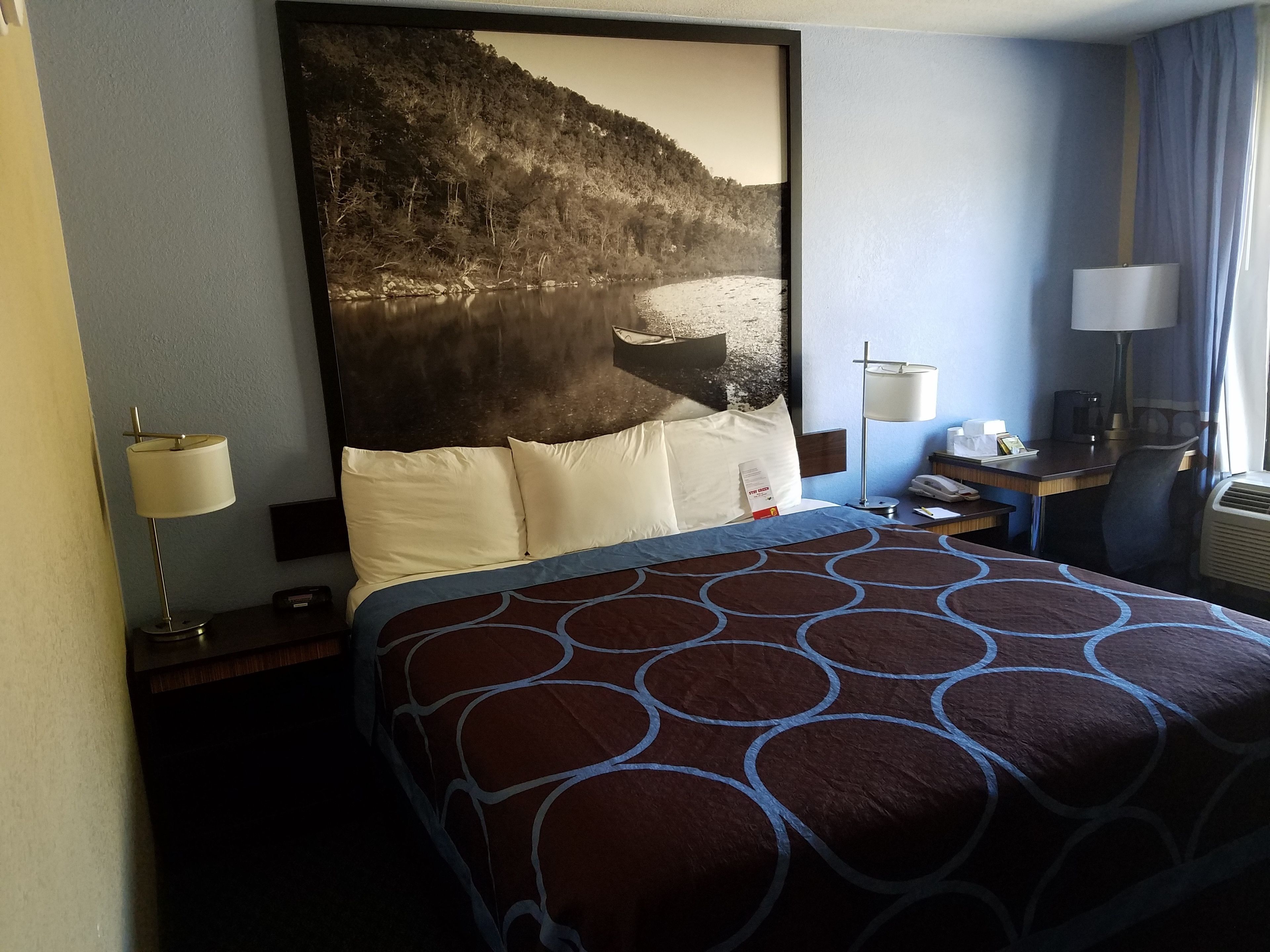 A hotel room with a large bed, a desk with a lamp, a chair, and a view of a forested area.