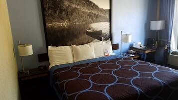 A hotel room with a large bed, a desk with a lamp, a chair, and a view of a forested area.