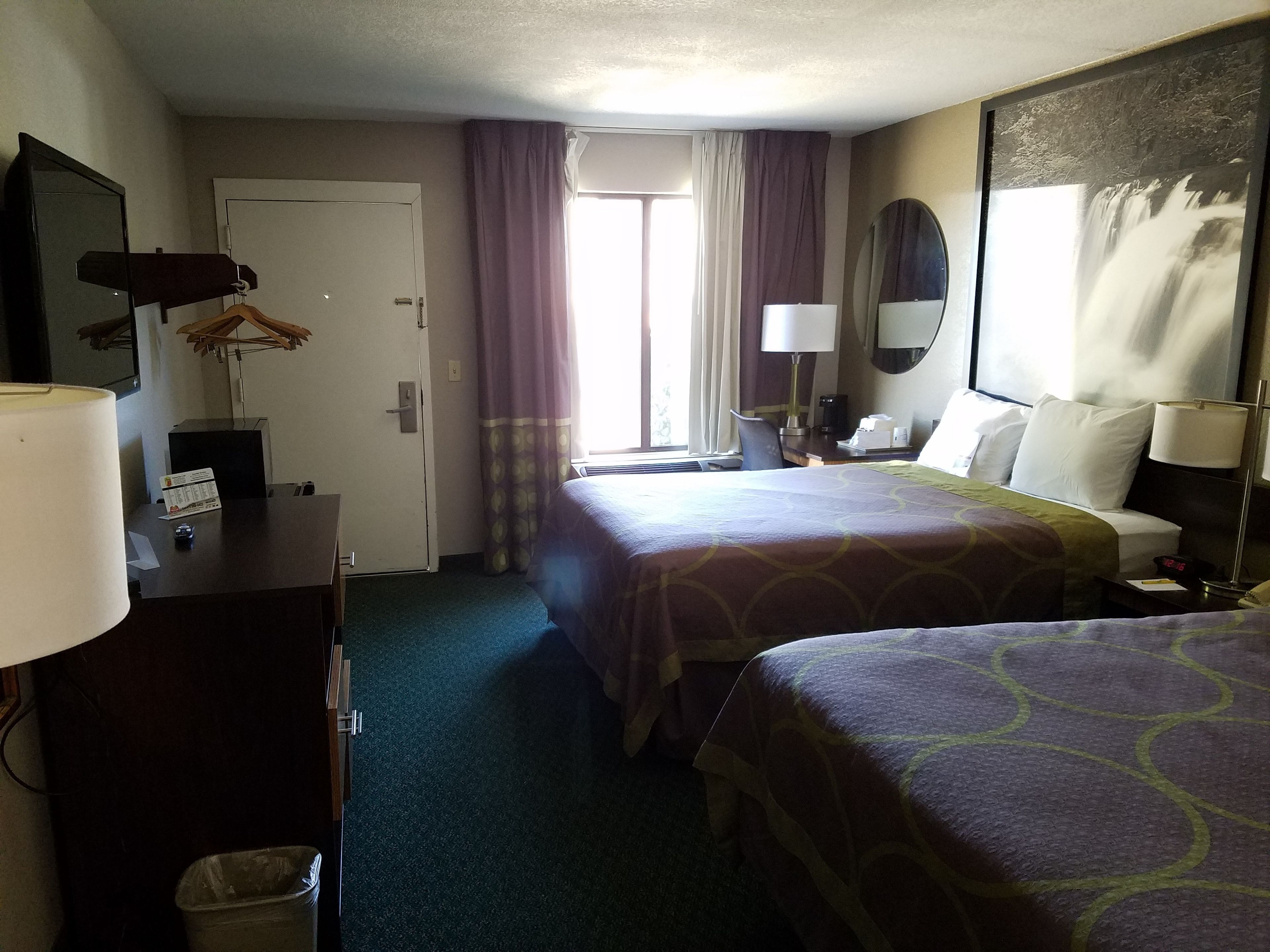 A hotel room with two beds, a desk with a lamp, a chair, a trash bin, and a window with curtains.