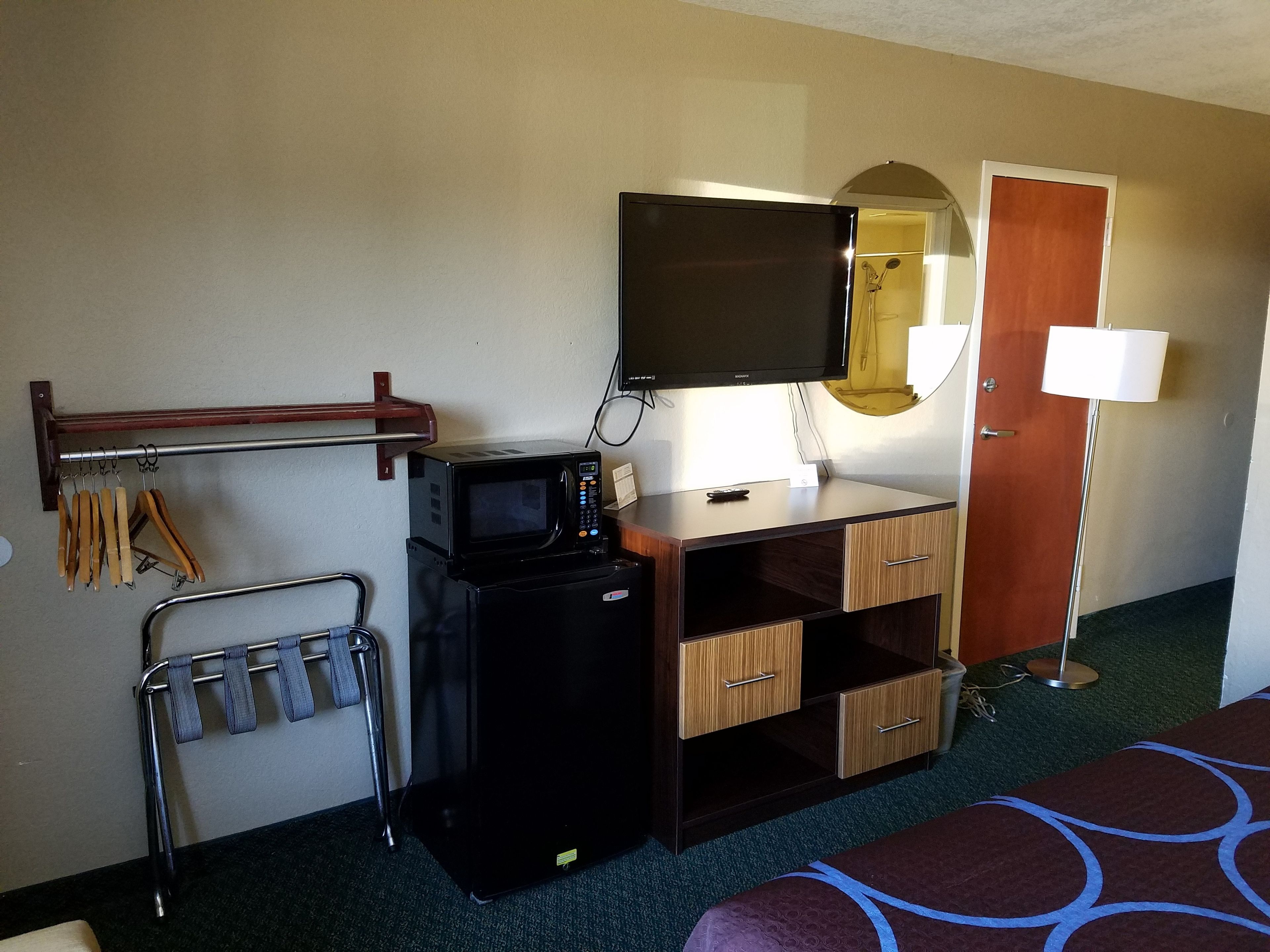 Ett kompakt hotellrum med ett litet kök, en platt-TV, ett skrivbord i trä, en spegel, en lampa och en säng med ett mönstrat överkast.