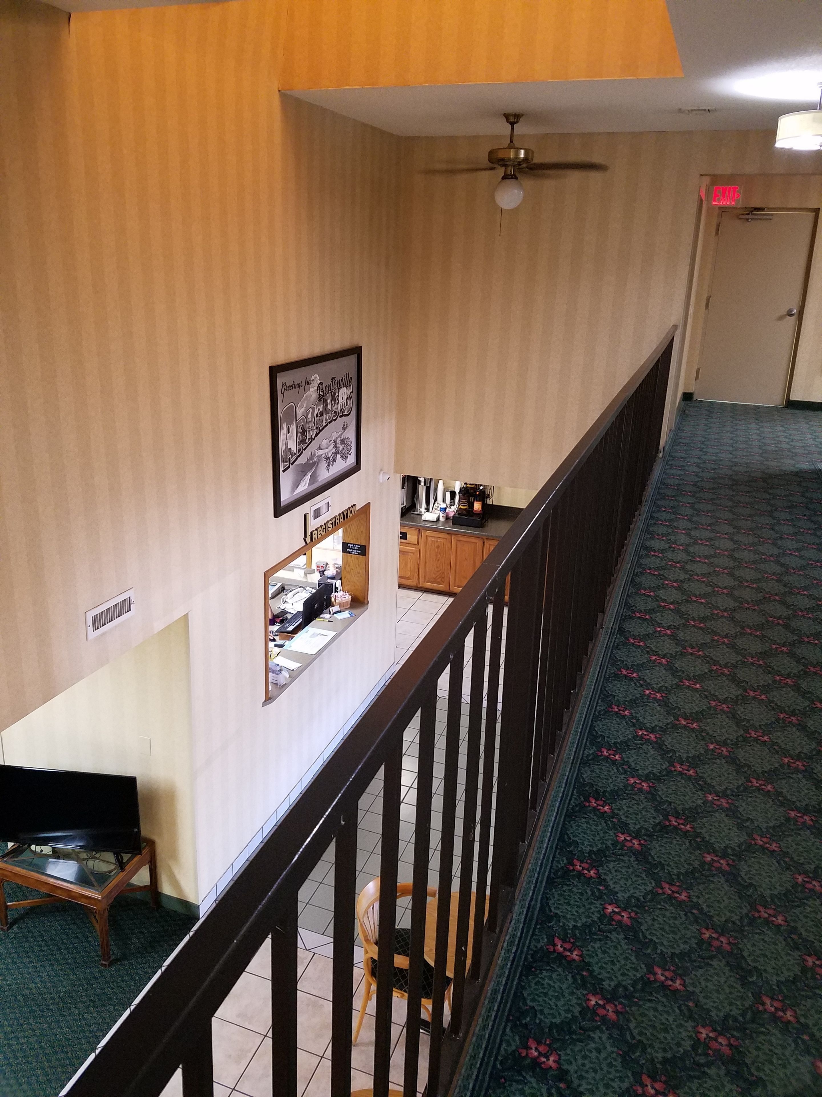 A hotel room with a desk, a chair, a ceiling fan, a registration desk, and a carpeted floor.