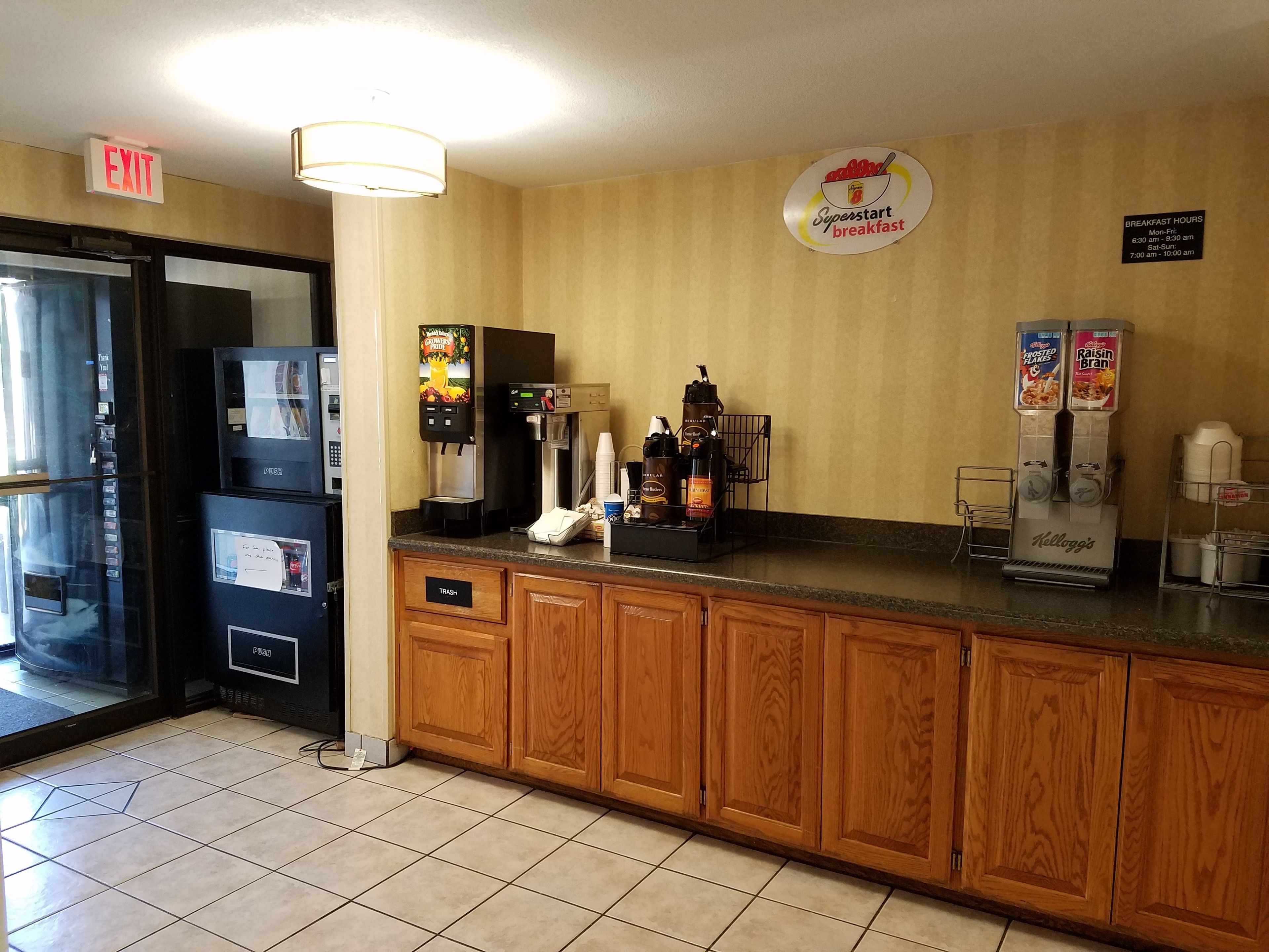 Una cocina de hotel con zona de desayuno tipo bufé, que incluye una máquina de café, dispensador de zumo y un refrigerador.