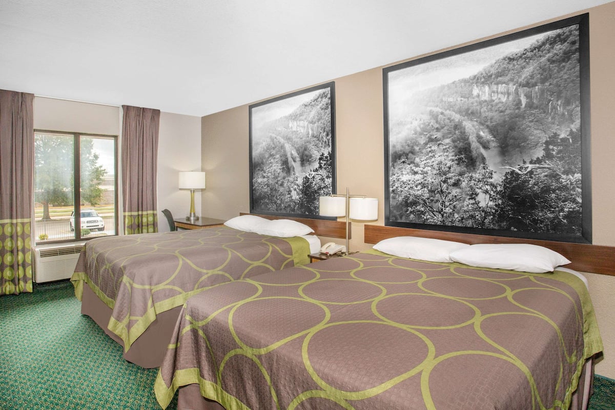 A hotel room with two beds, a large window, and two large landscape paintings on the wall.