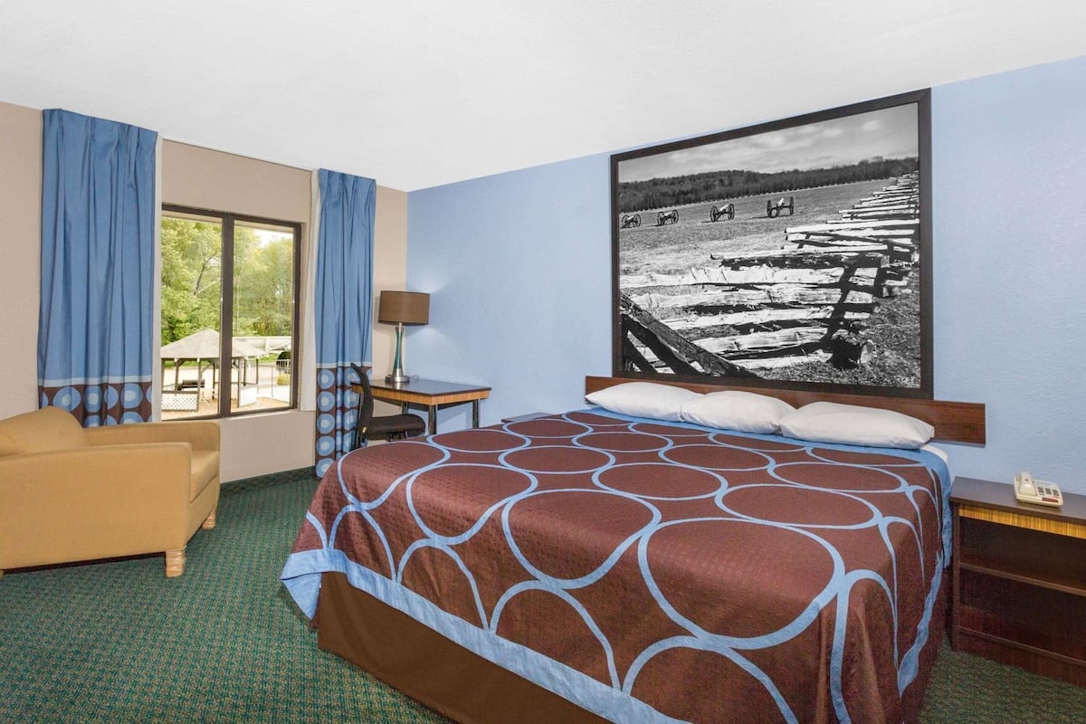 A hotel room with a bed, a desk, a chair, a window with curtains, and a large framed picture on the wall.