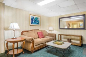 A hotel room with a beige sofa, a small round table with a lamp, a coffee table with a decorative bowl, a large mirror, and a framed abstract painting on the wall. - Super 8 by Wyndham Bentonville (Bentonville)