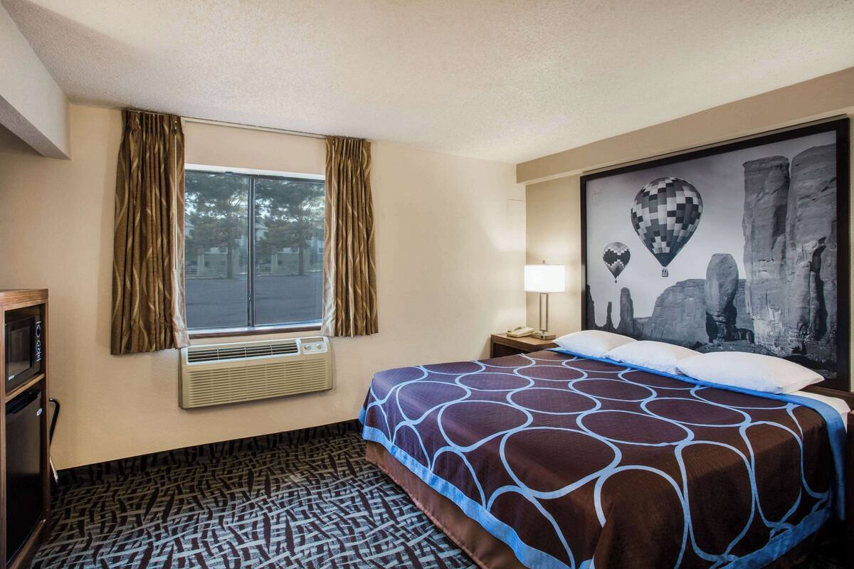 Habitación de hotel con una cama grande, una ventana con cortinas, un aire acondicionado de pared, una mesita de noche con lámpara y un cuadro en la pared.