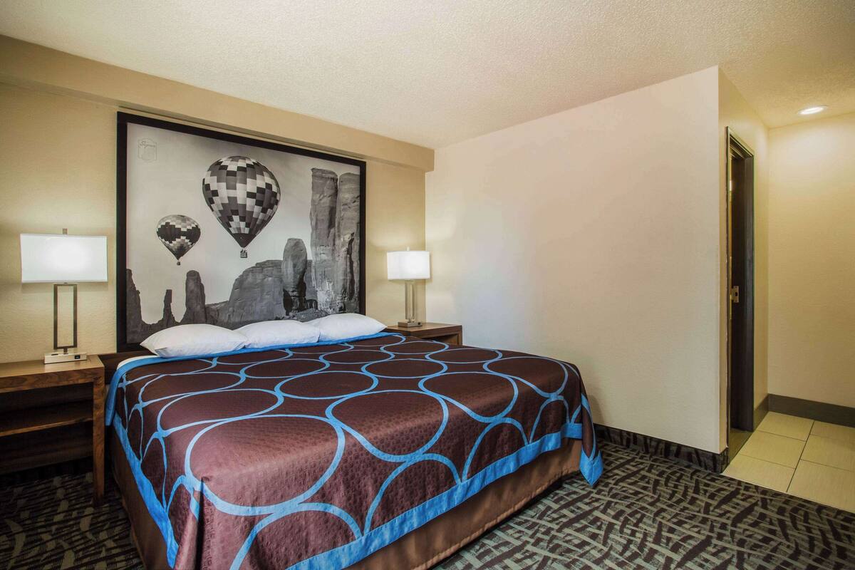 Habitación de hotel con una cama grande, dos mesitas de noche, una lámpara de pared y una obra de arte con una vista urbana enmarcada.