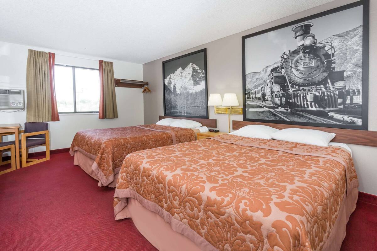 A hotel room with two beds, a desk, a chair, a TV, and a large framed picture of a train on the wall.