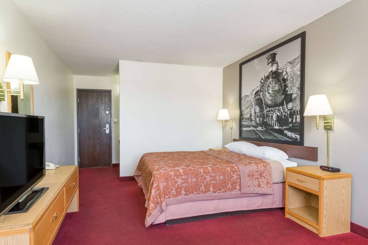 A hotel room with a bed, a TV, a wooden nightstand, a lamp, and a framed picture on the wall.