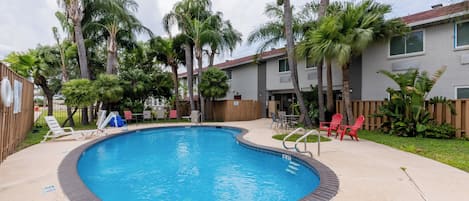 Piscina con borde de ladrillo, rodeada de palmeras y sillas rojas.