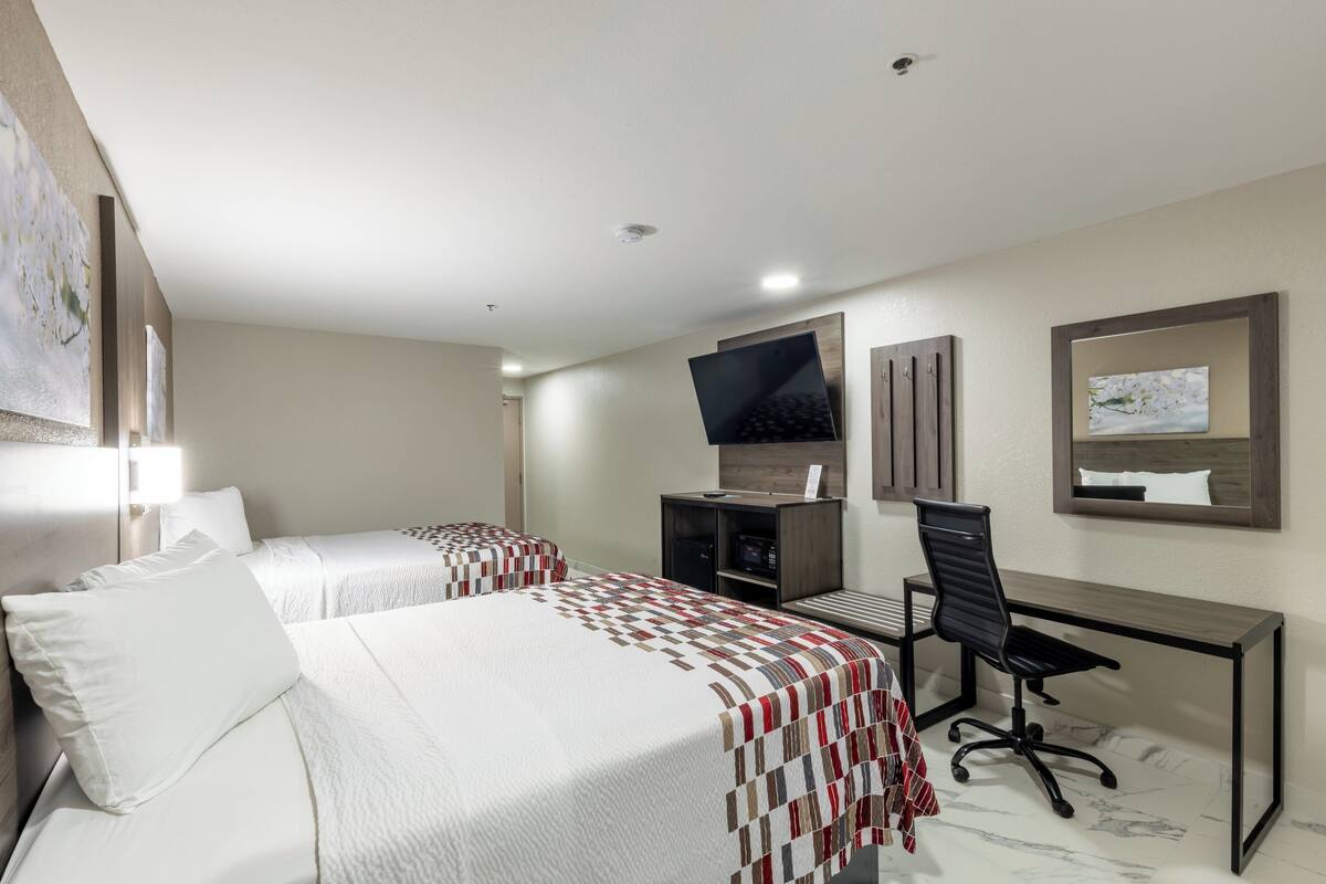 A hotel room with two beds, a desk, a chair, a TV, and a painting on the wall.